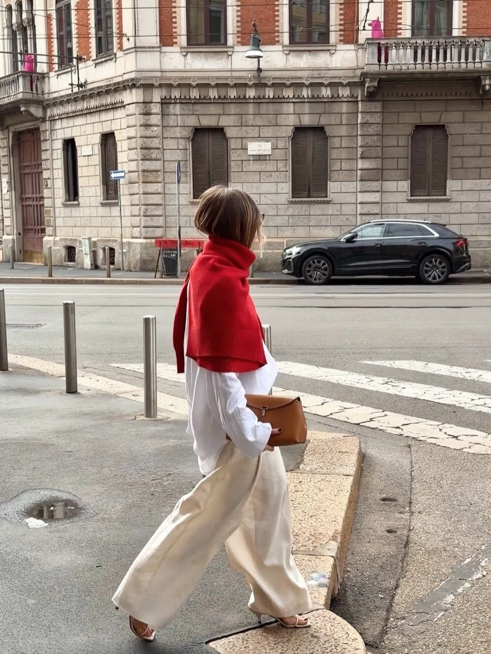 Person walking on a city sidewalk wearing a red shoulder-draped sweater, white shirt, and cream wide-leg trousers.