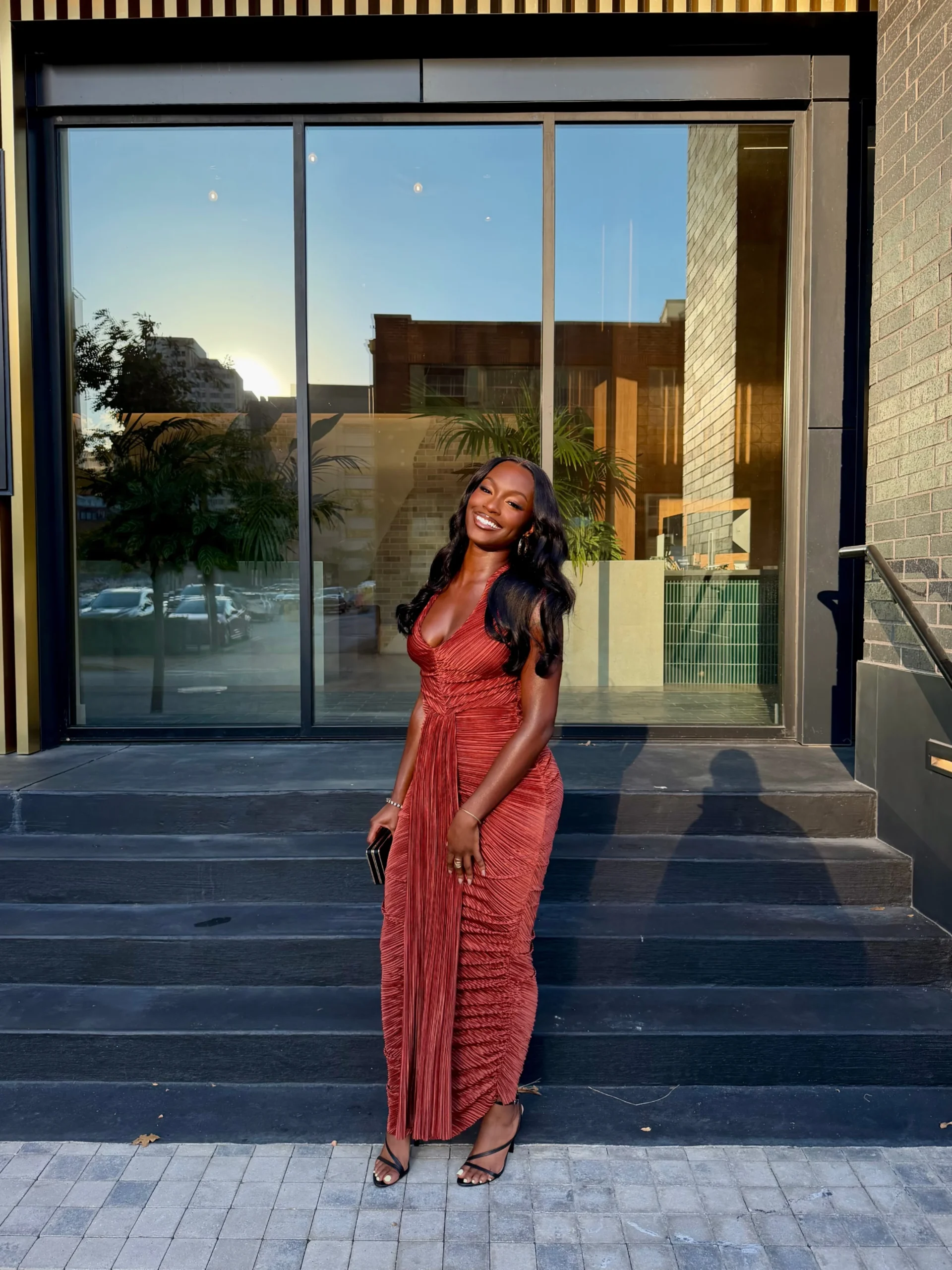 Woman wearing a rust-red ruched sleeveless maxi dress with black strappy heeled sandals.