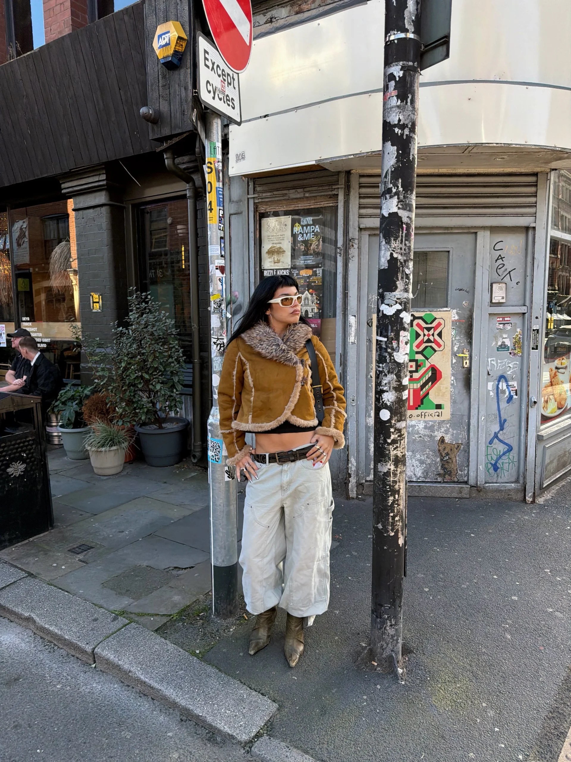 Cropped tan shearling jacket with wide-leg trousers, dark belt, and pointed olive-brown ankle boots.