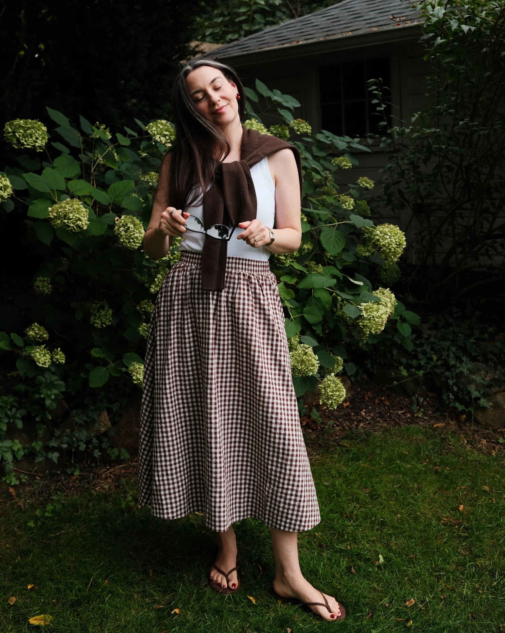 White sleeveless top tucked into a brown gingham skirt with a draped brown sweater and sandals.