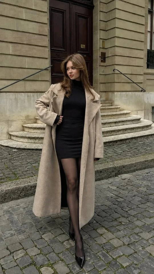 Woman in a beige overcoat, black highneck mini dress, sheer tights, and patent stilettos.