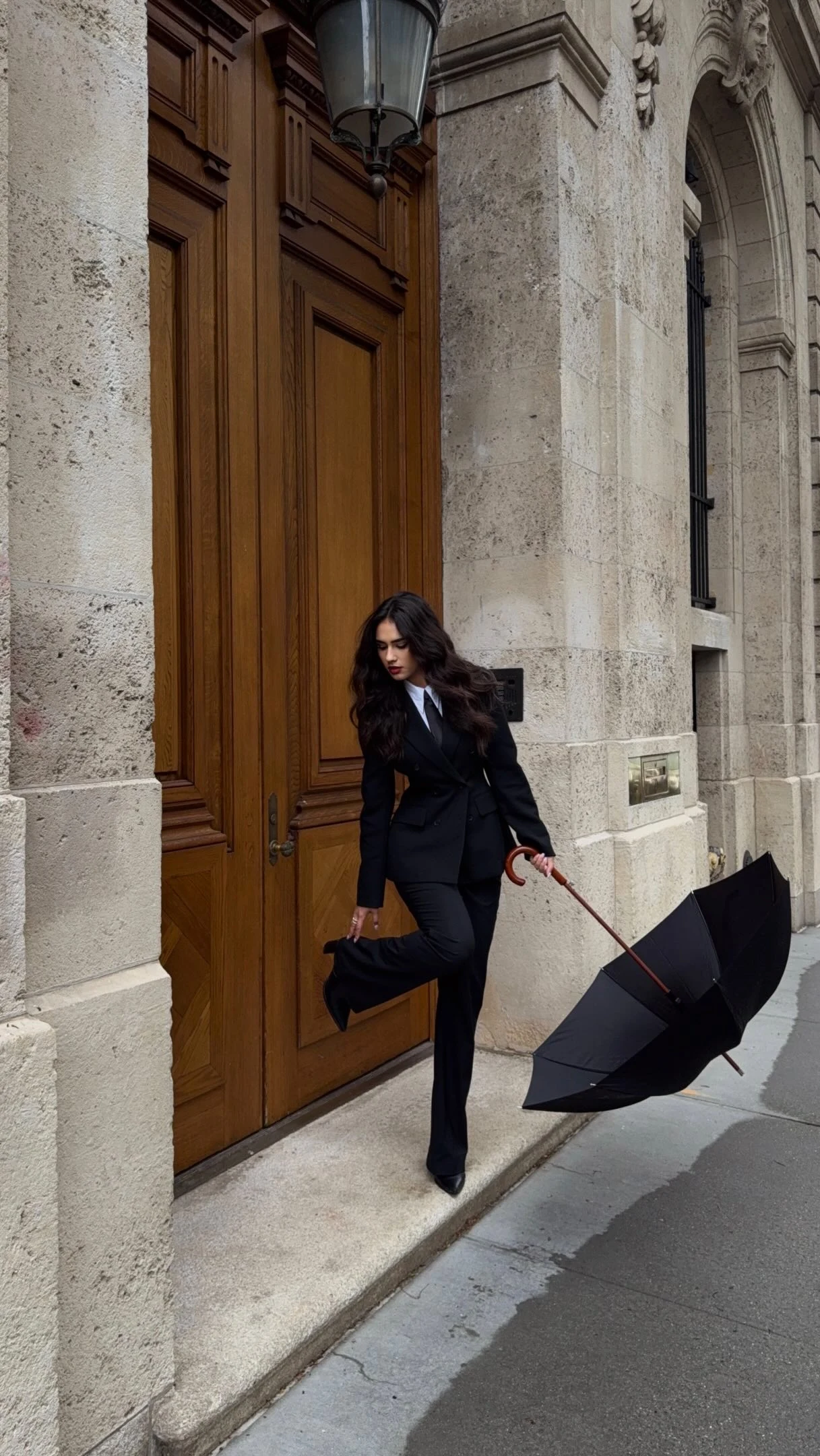 A fitted black blazer with notched lapels and matching straight-leg trousers forms a coordinated suit. A white collared shirt and narrow black tie are visible underneath. The outfit is paired with pointed black shoes. The person holds an open black umbrella with a curved brown handle.