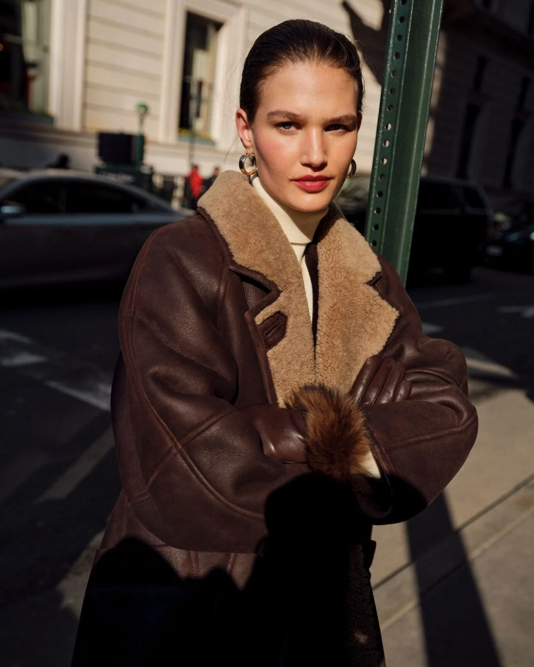 Oversized dark brown shearling-collar jacket over a cream turtleneck with brown gloves and gold hoops.