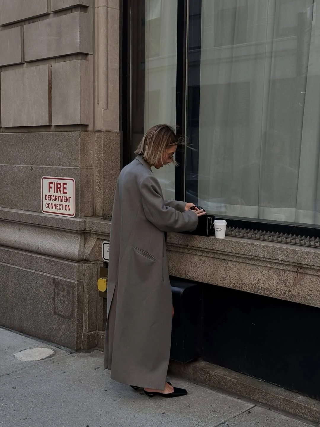 Person in an oversized grey double-breasted long coat, brown shirt, blue jeans, and black structured handbag.