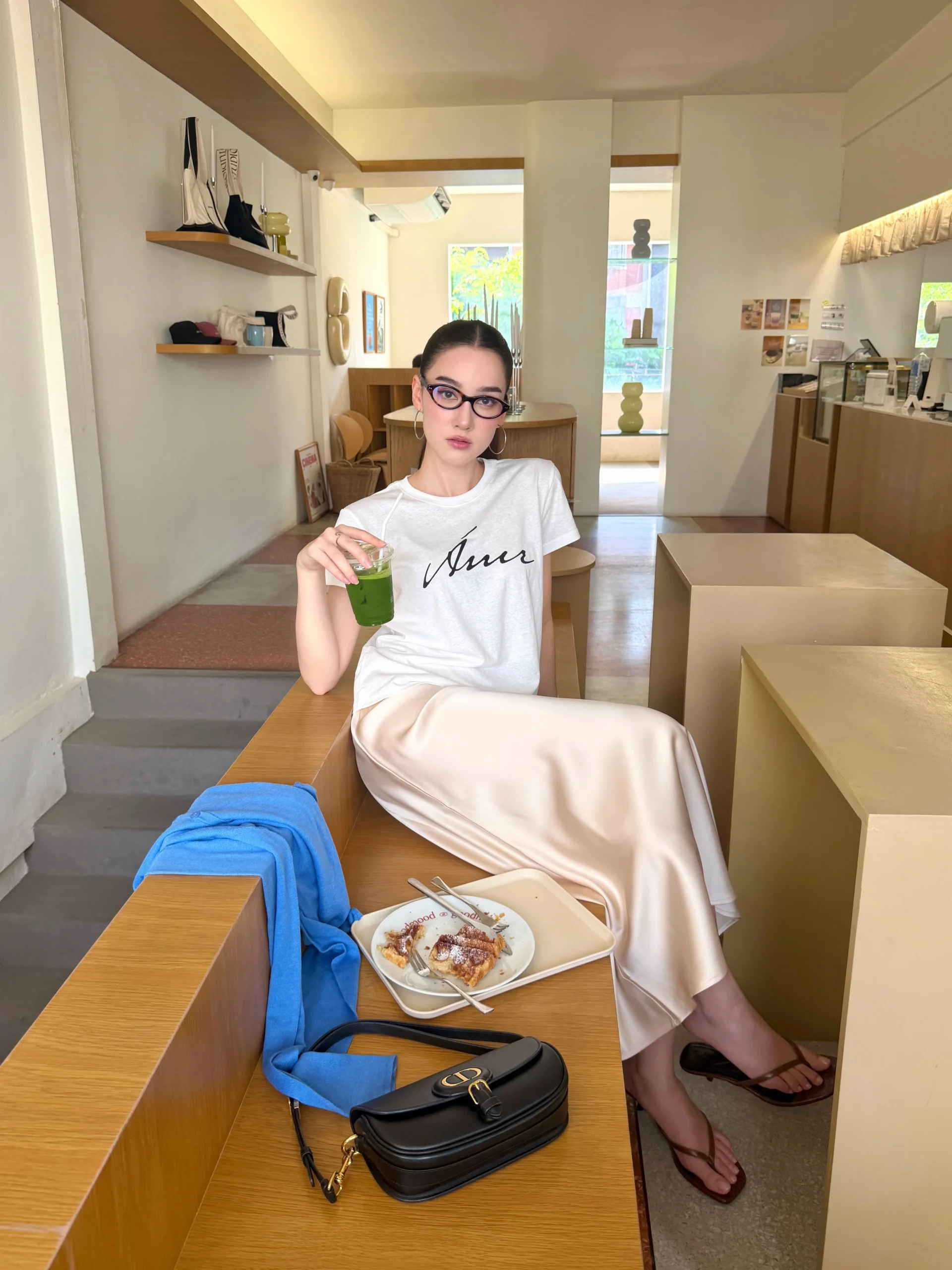 Woman in a graphic tee, cream satin skirt, blue tied sweater, and brown sandals.