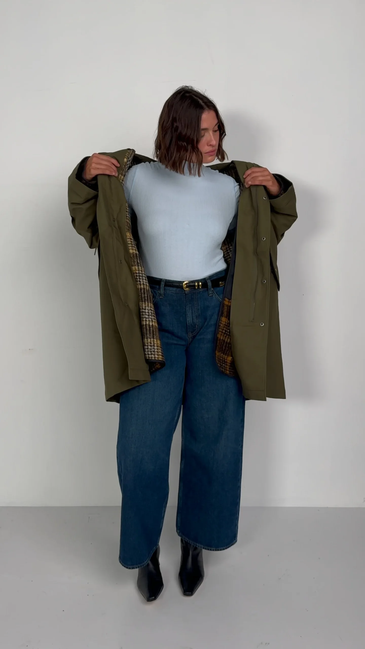 Open olive green checked coat over a pale blue sweater, wide-leg jeans, and black ankle boots.