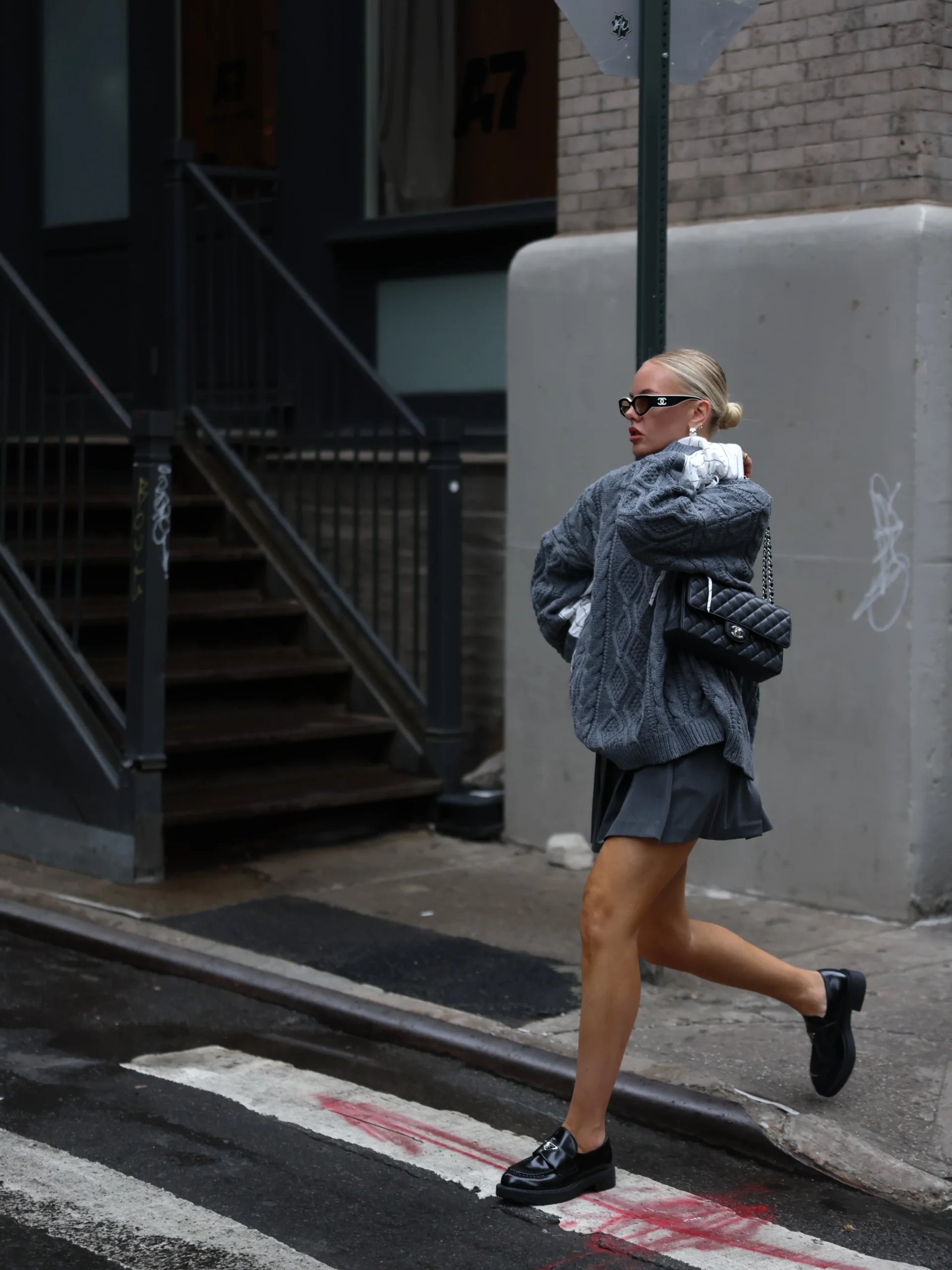 Oversized gray cable-knit sweater layered over white pinstriped shirt with ruched cuffs, short dark gray pleated mini skirt, black platform loafers, and black quilted shoulder bag.