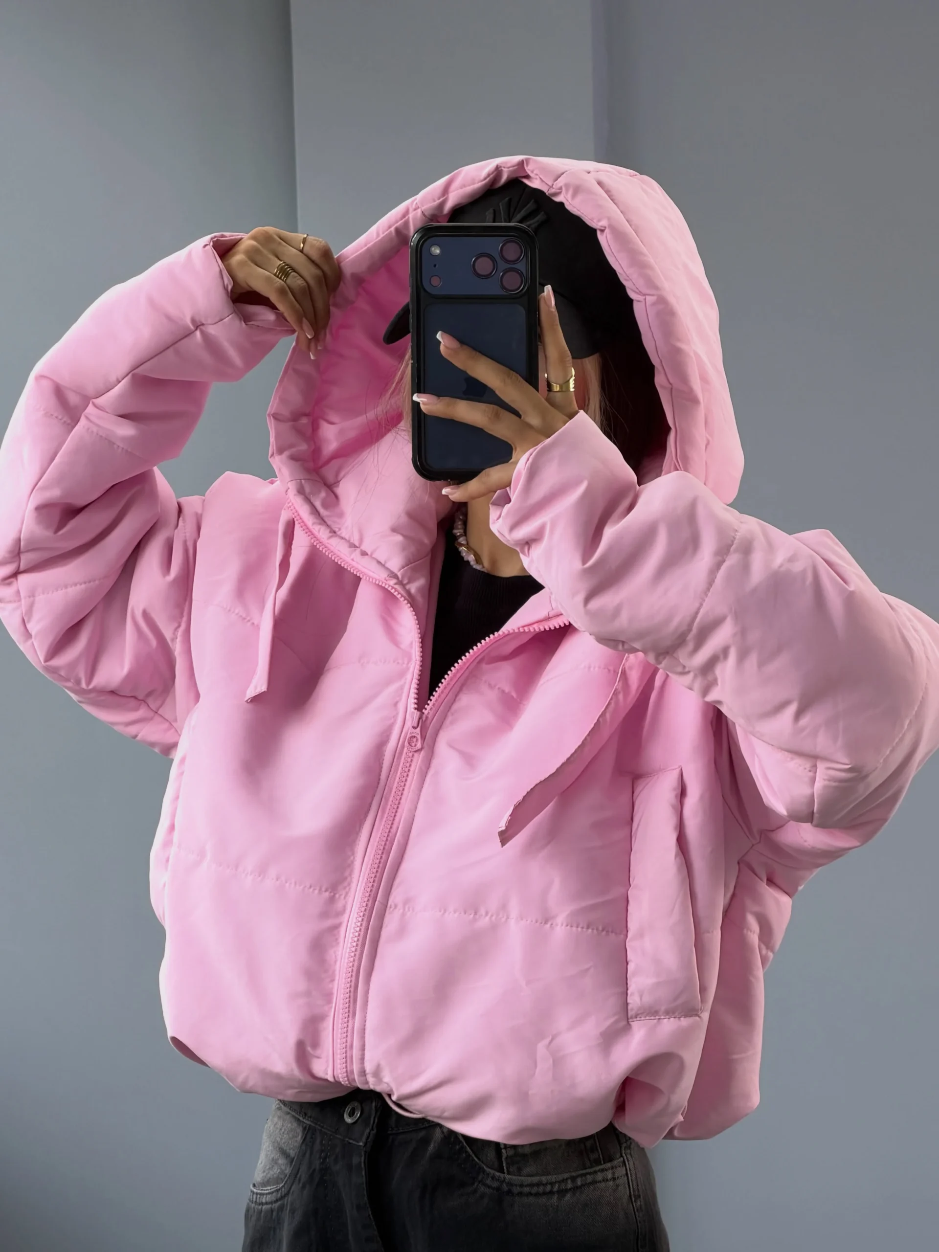 Bright pink hooded puffer jacket with hood up worn over a black top and dark charcoal jeans.