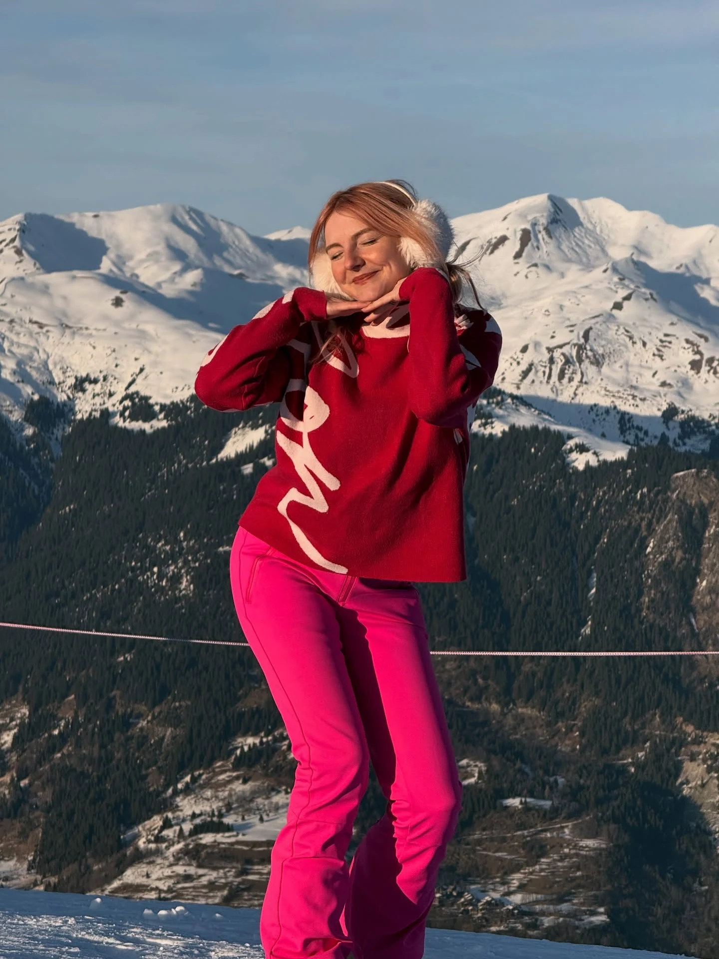 Woman in a red knit sweater, bright pink ski pants, and white fluffy earmuffs in a winter setting.