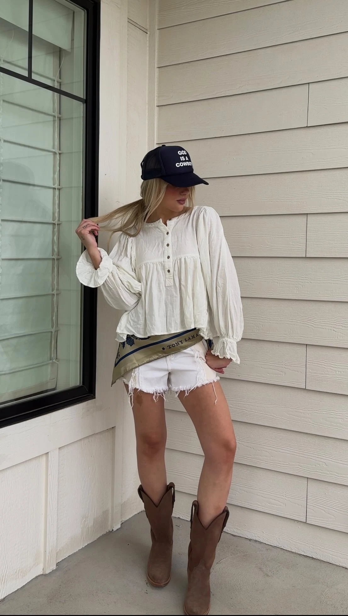 Woman in an olive sheer ruffled blouse, distressed denim shorts, and brown suede cowboy boots.