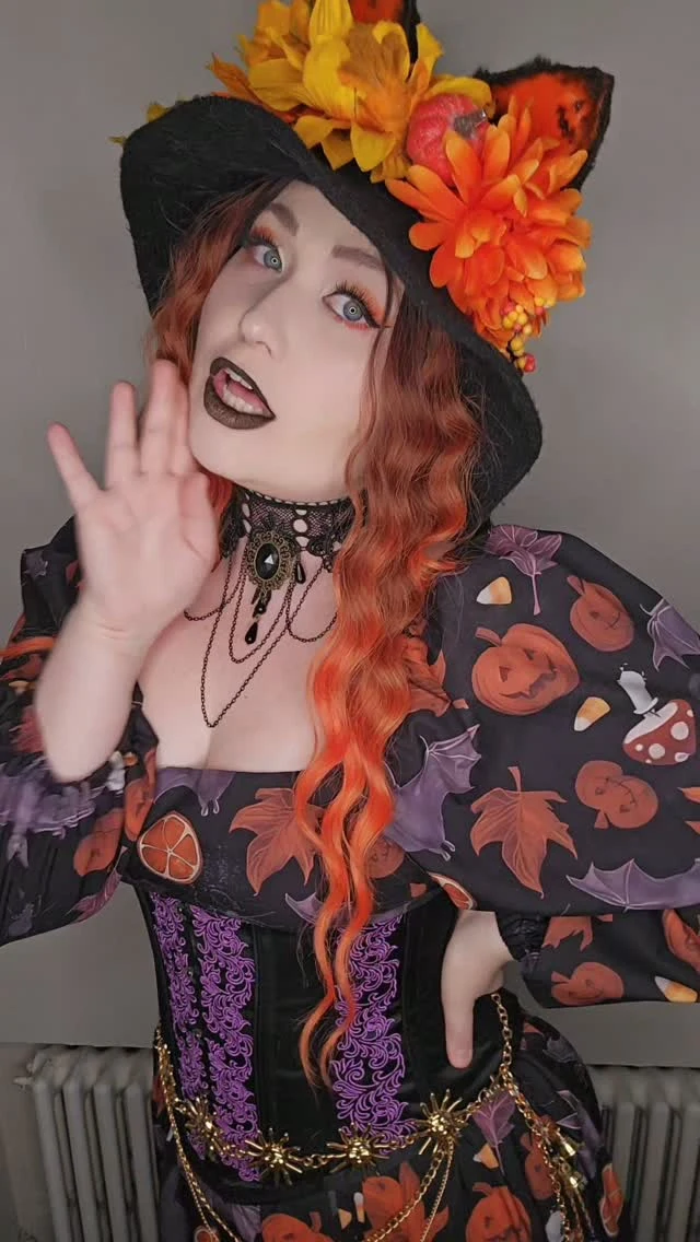 Black wide-brimmed hat with orange and yellow flowers and pumpkins, black dress with bats leaves print over black corset with purple lace-up