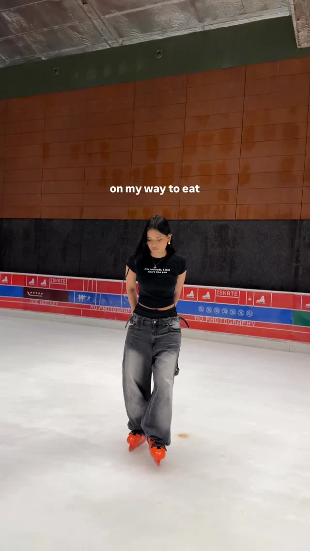 Person in black cropped tee and wide-leg gray jeans with orange-red ice skates on an indoor rink.