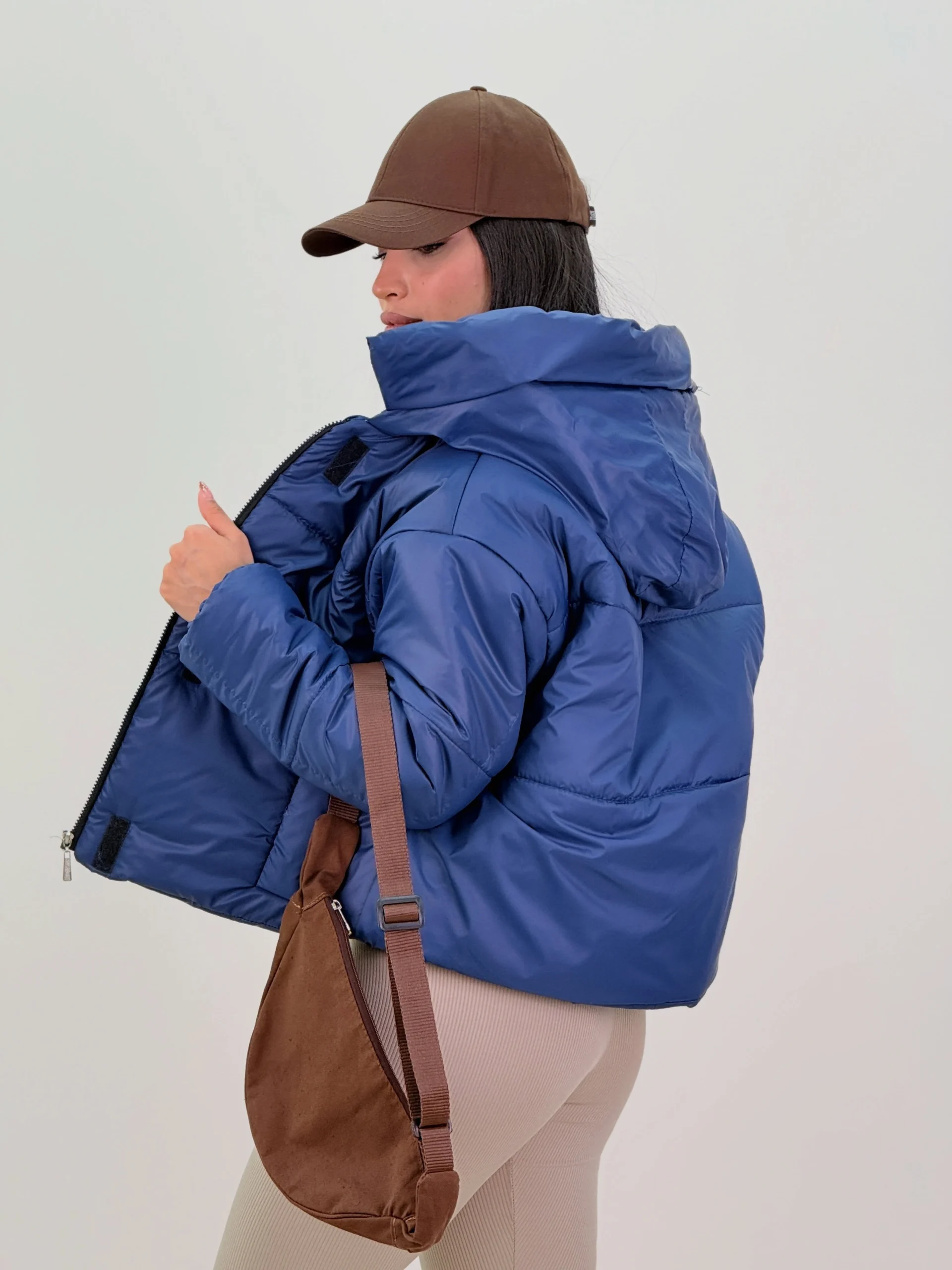 Bright blue cropped hooded puffer jacket with beige ribbed leggings, brown cap, and brown shoulder bag.