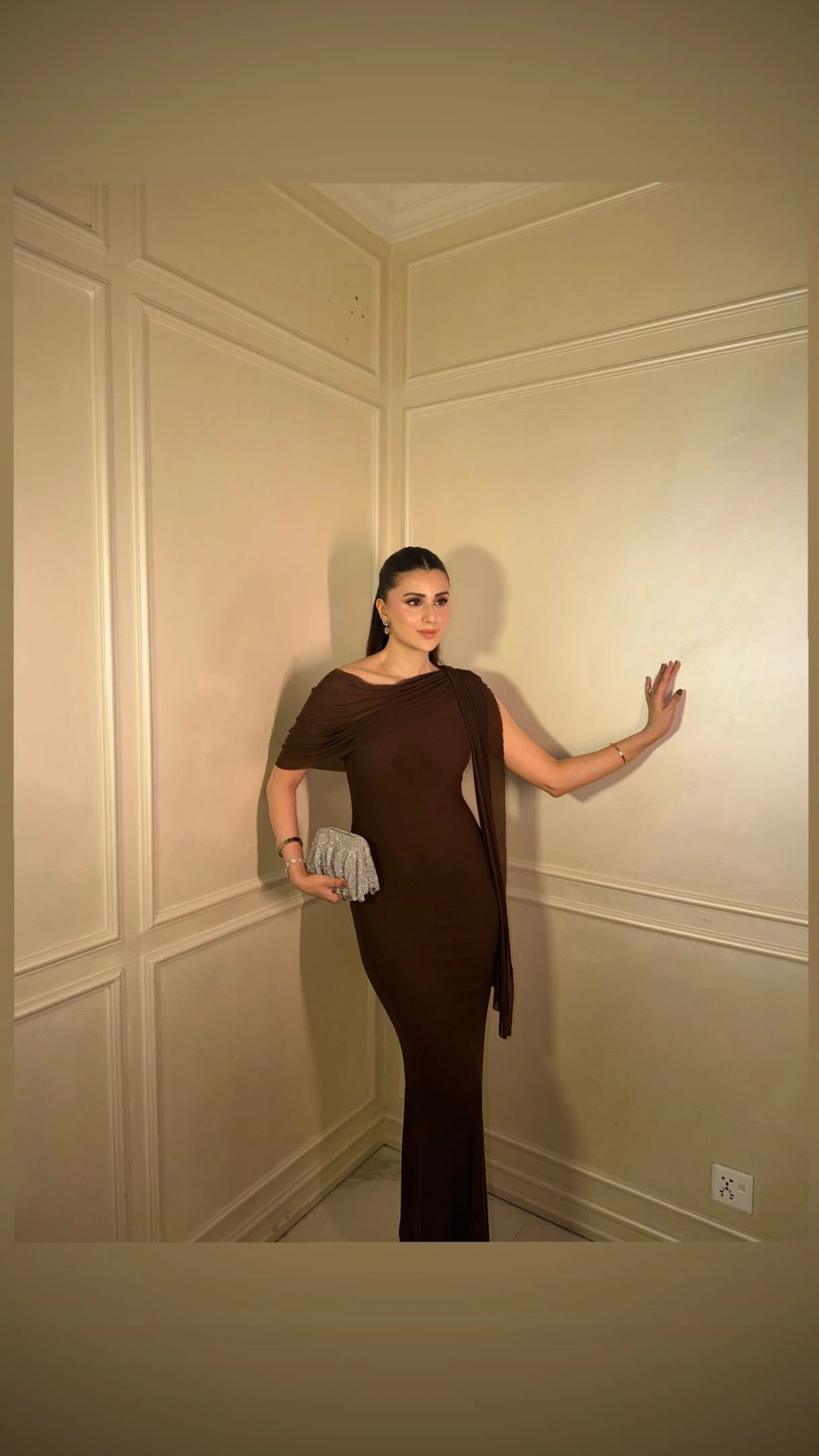 Woman in a dark brown draped floor-length dress holding a small silver textured clutch.