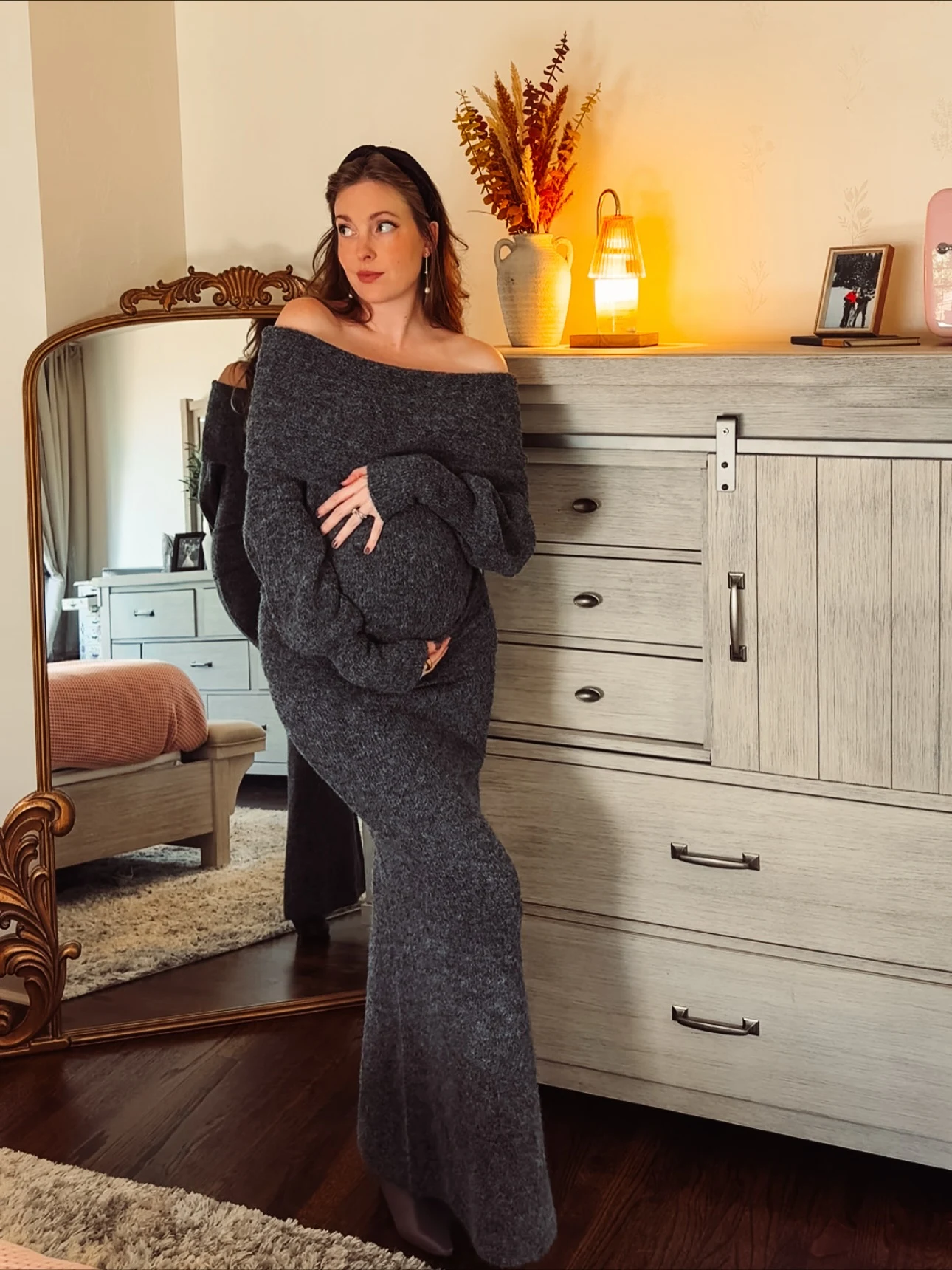 Pregnant woman in a floor-length charcoal off-shoulder knit dress standing beside a light wood dresser.
