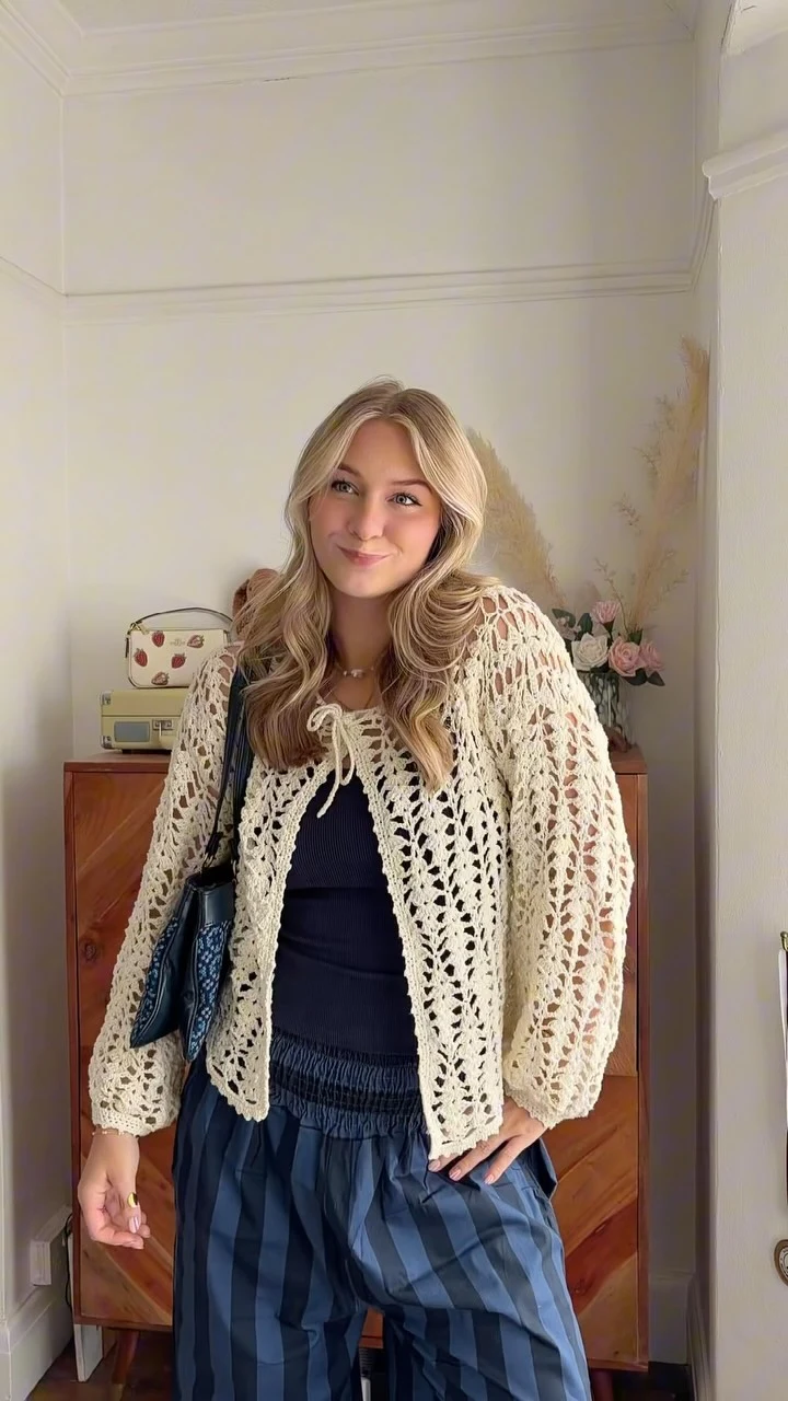 Cream open-knit cardigan with tie neckline layered over navy top and blue-gray check trousers.