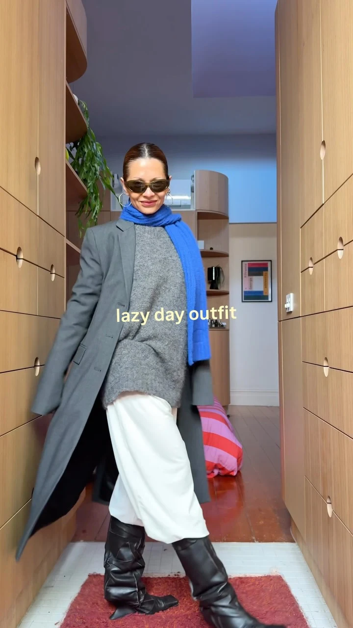 Woman in an oversized gray sweater, white drawstring pants, knee-high boots, and a bright blue scarf.