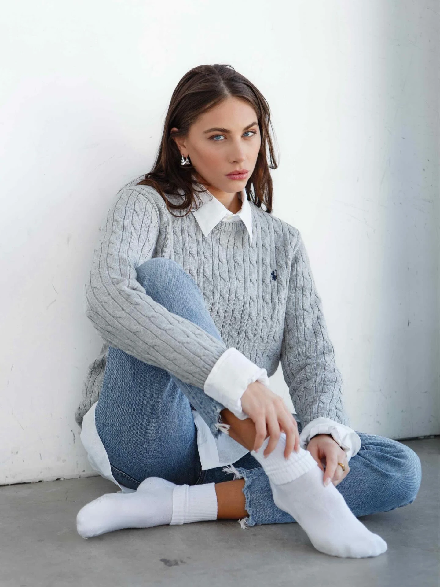 Light grey polo cable-knit sweater layered over white button-down shirt with rolled cuffs, light blue distressed straight jeans, white crew socks