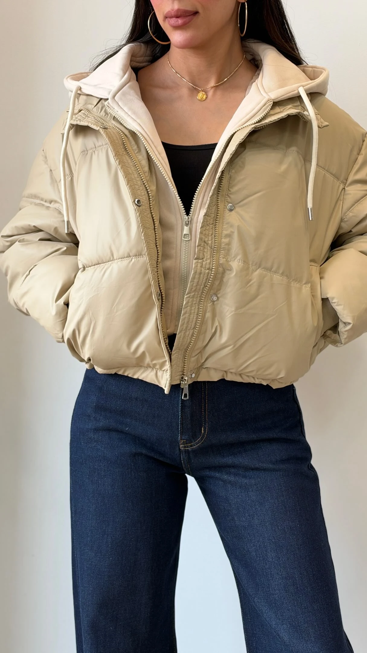 Light beige cropped puffer jacket with white hood worn over a black top and dark blue jeans.