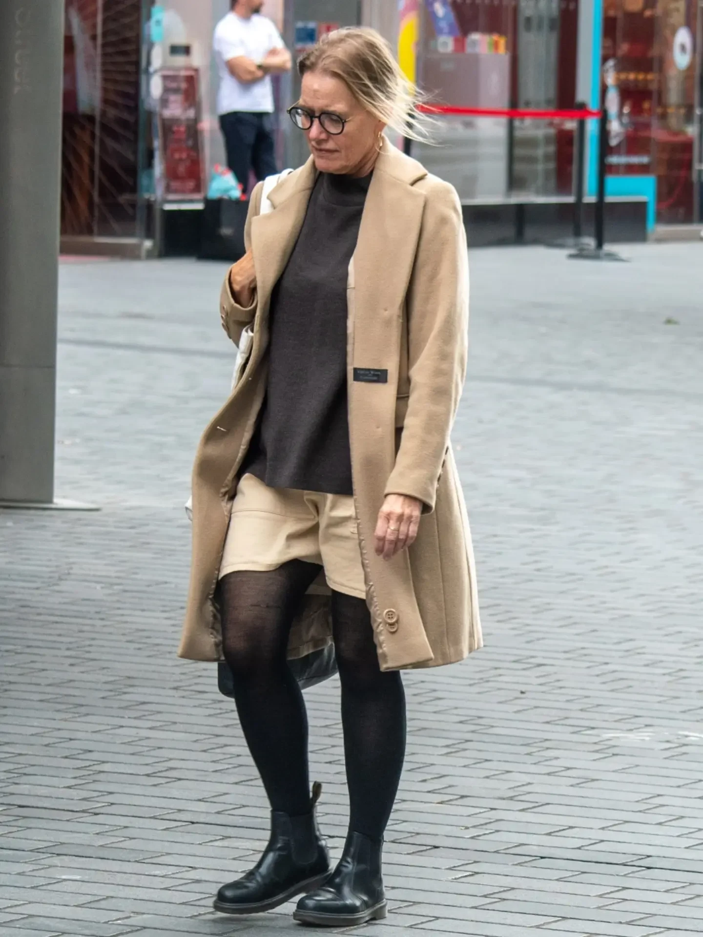 Long camel coat over beige shorts, sheer black tights, and chunky black lace-up ankle boots outdoors.