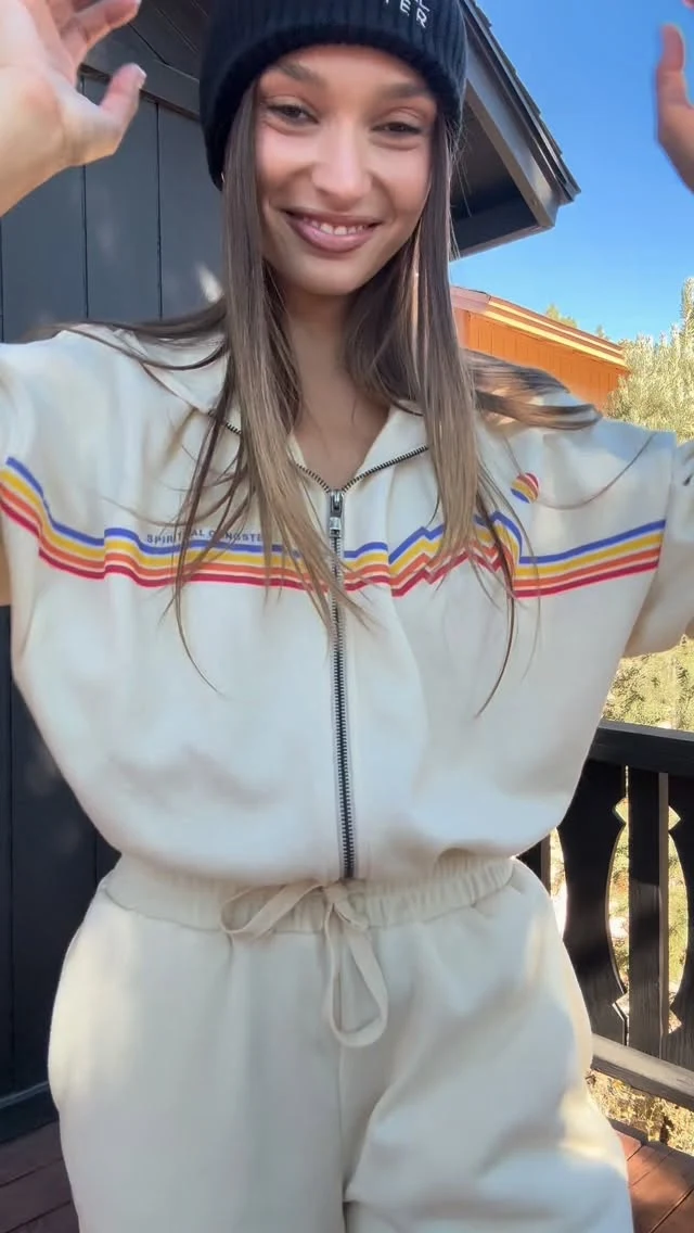 Cream matching track jacket and joggers with colorful chest stripes and a black knit beanie.