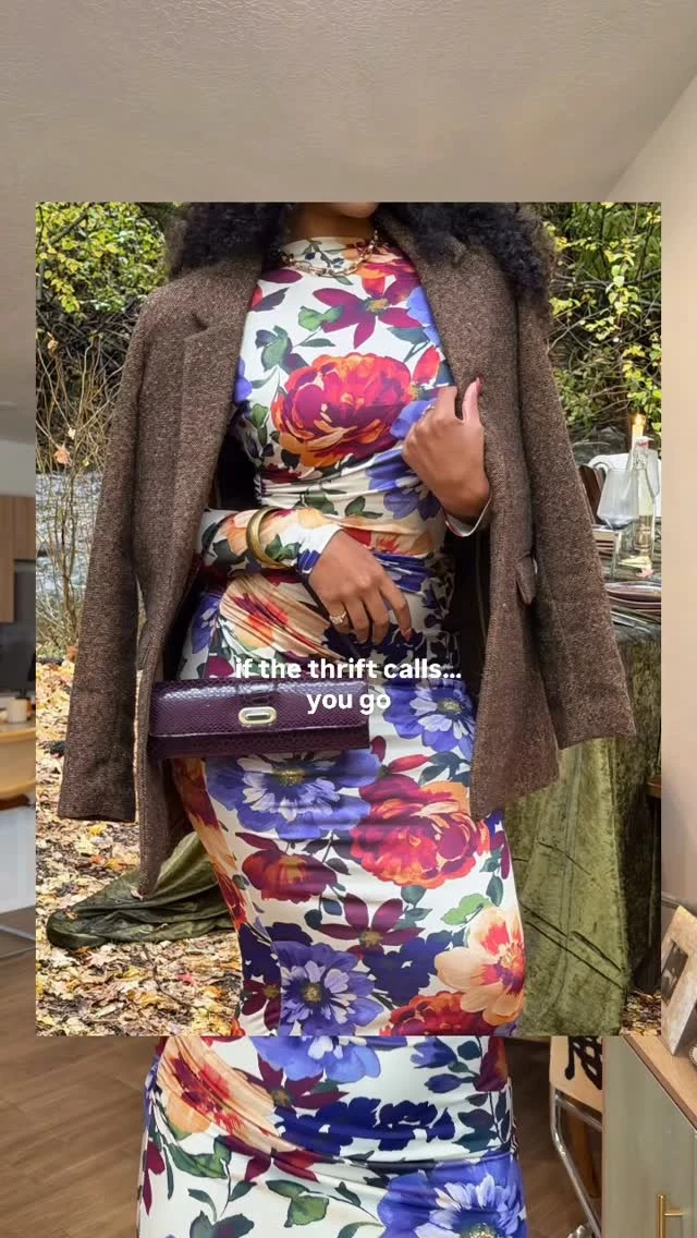 Woman in a white floral ankle-length dress layered under an open brown coat, holding a purple bag.