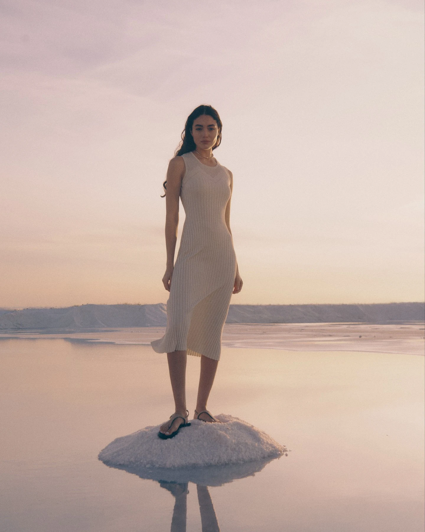 Ivory sleeveless ribbed midi dress with V-neckline, hem lifted by breeze, and dark flat sandals.