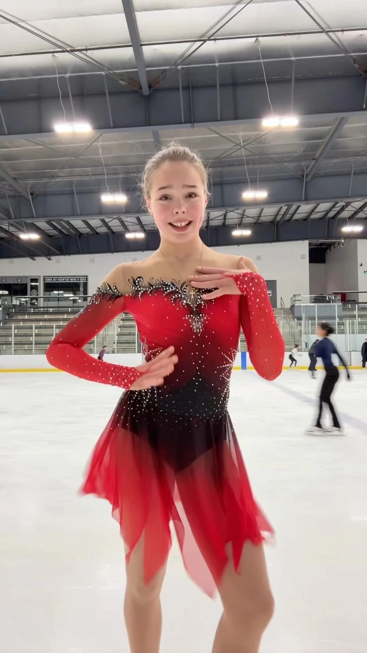 Person in fitted red and black embellished one-shoulder dress with layered skirt on an indoor ice rink.