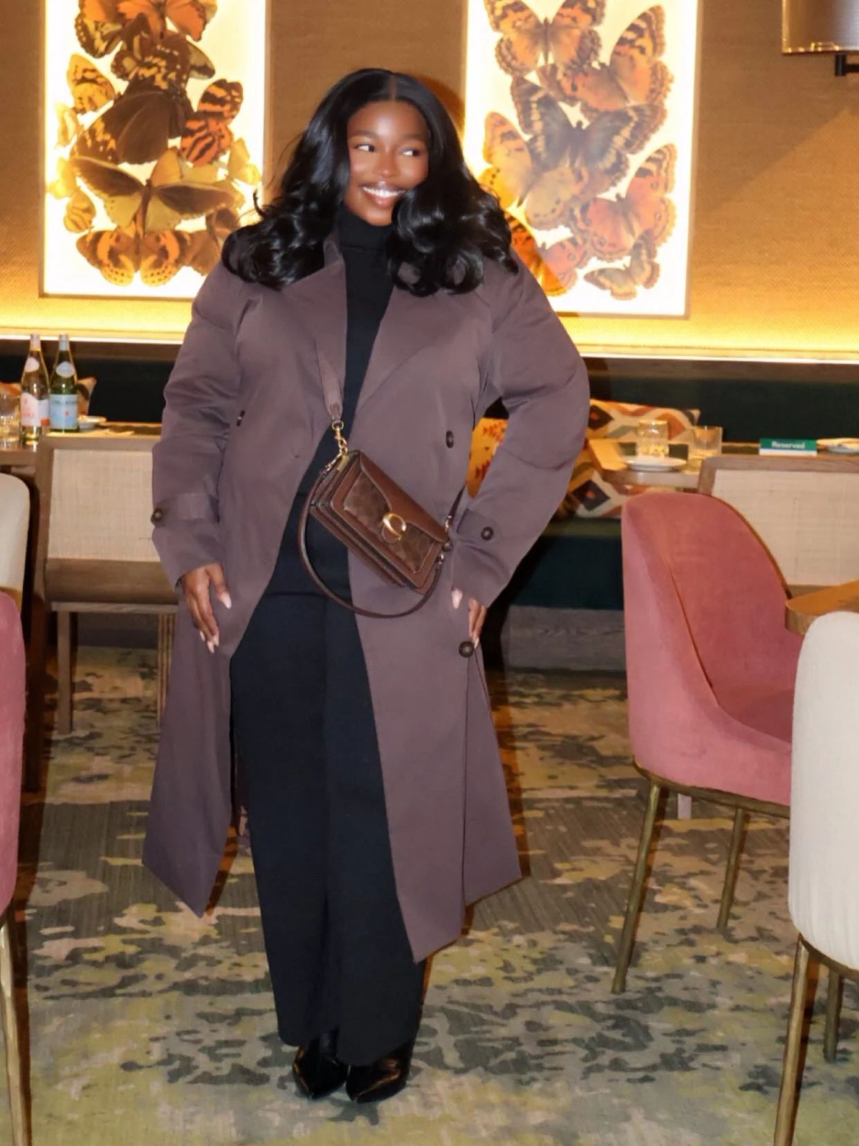 Woman in a taupe trench coat, black turtleneck, wide-leg trousers, ankle boots, and a monogram crossbody bag.