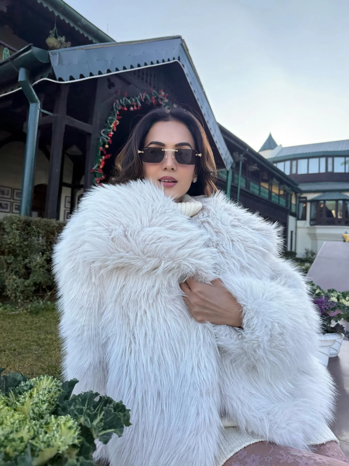Pregnant woman wrapped in a white fluffy coat over a cream turtleneck wearing dark oversized sunglasses.