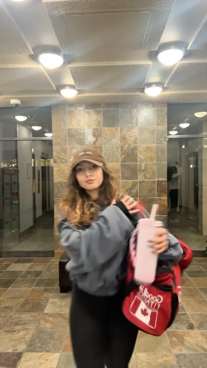 Person in a loose gray sweatshirt, black leggings, and tan cap holding a pink bottle.