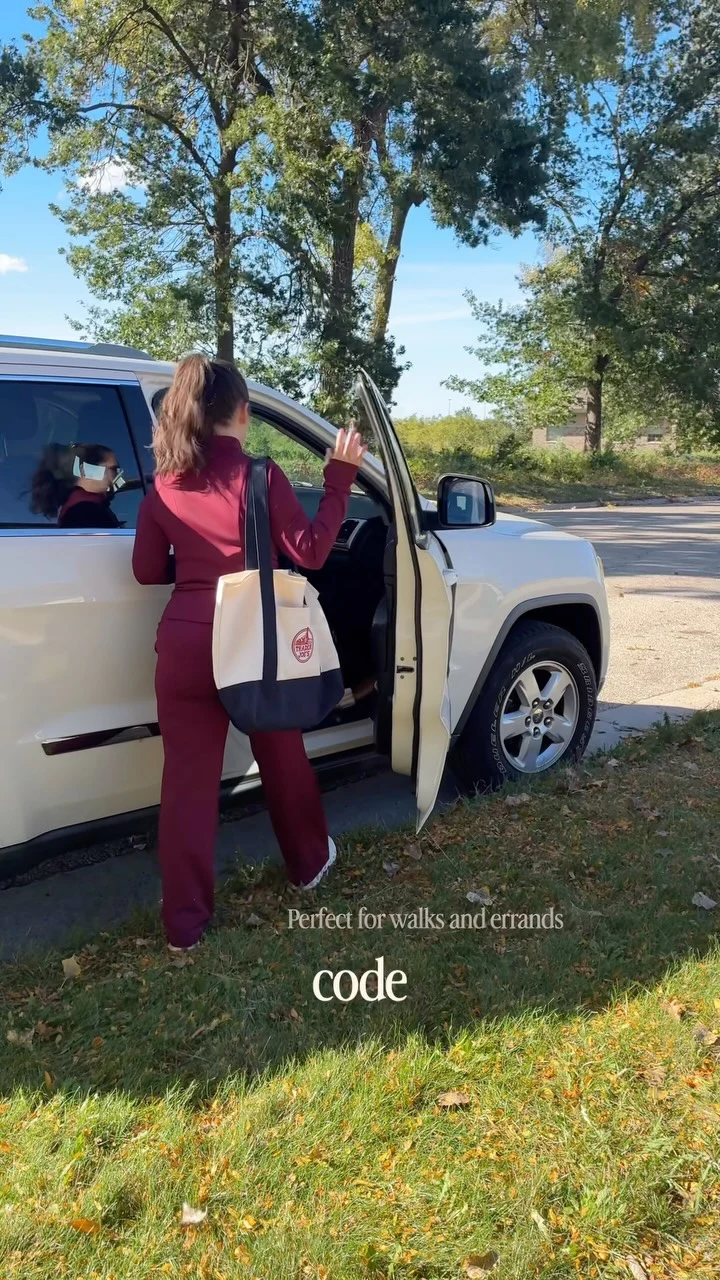 Person in a matching burgundy zip top and straight-leg pants carrying a cream-and-navy tote bag.